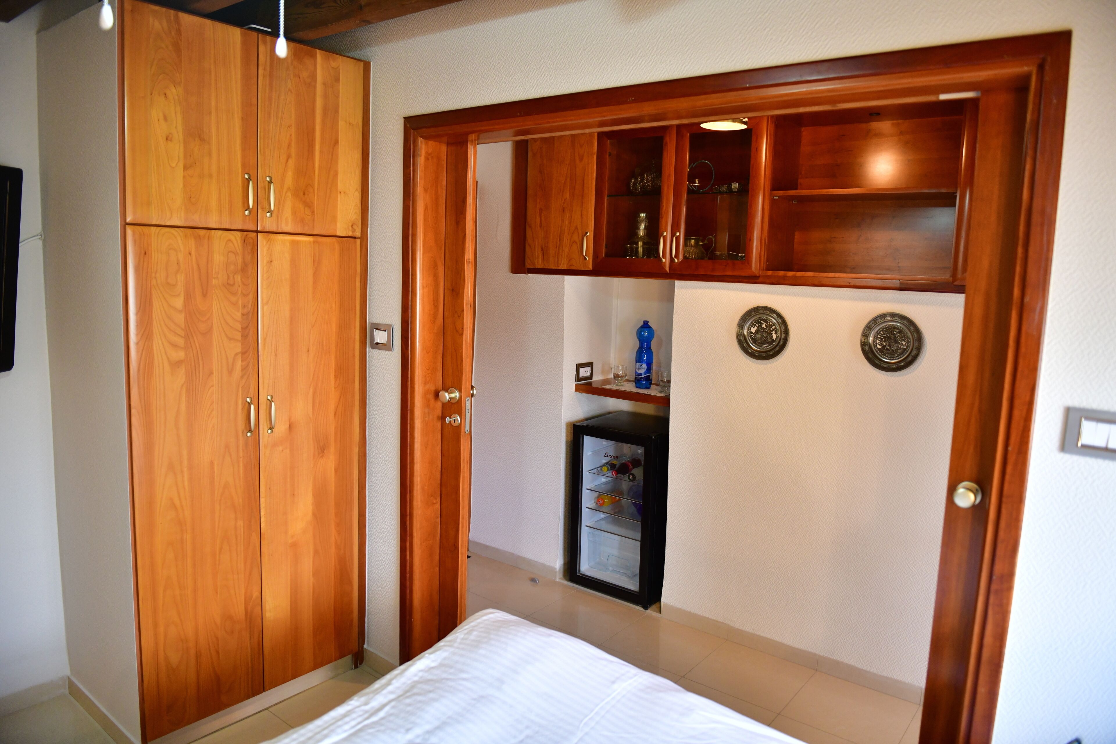 A bedroom with a wooden wardrobe, a bed, and a built-in mini-fridge.