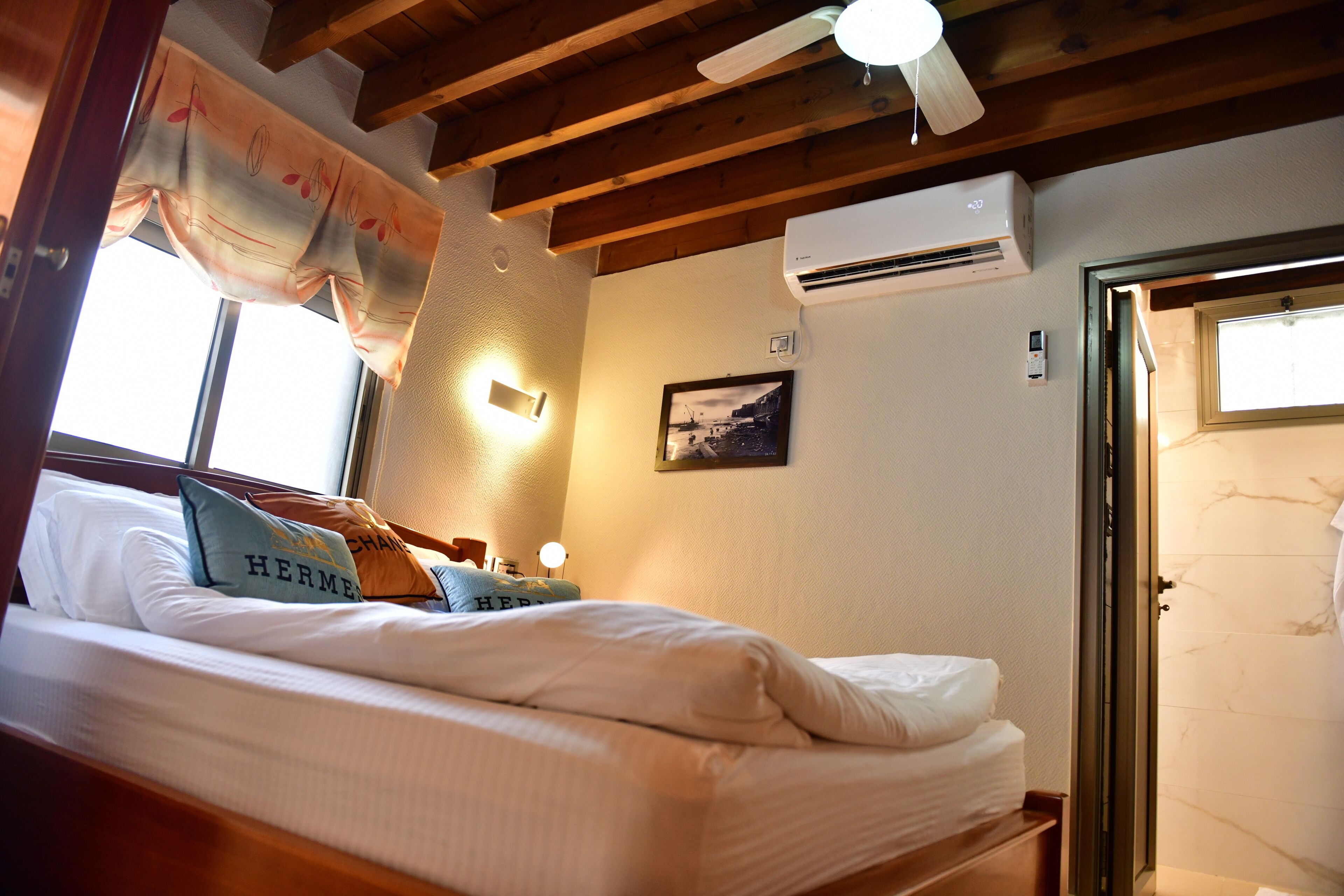 A hotel room with a bed, a ceiling fan, an air conditioning unit, a framed picture on the wall, and a door leading to a bathroom.