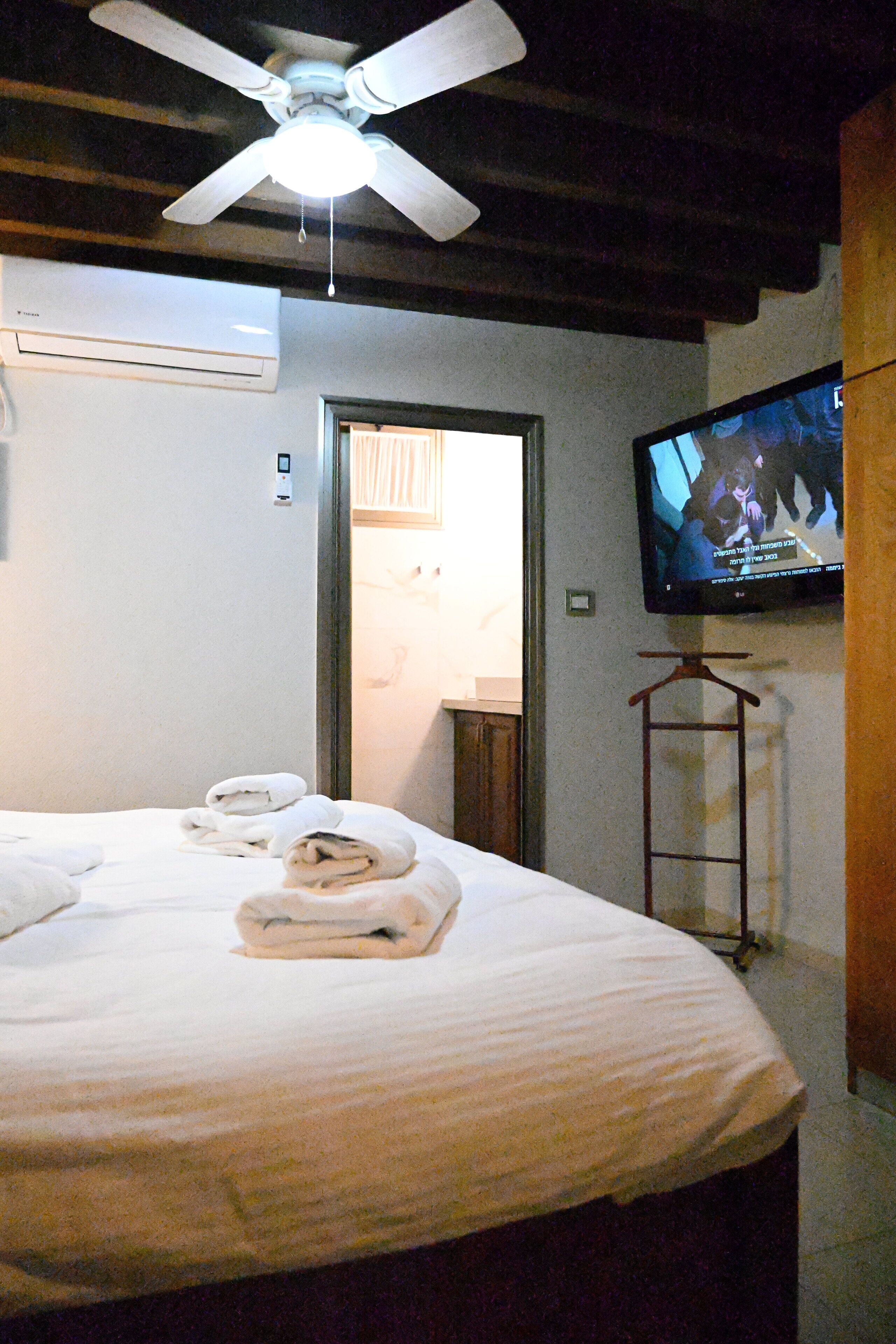 Quarto de hotel com cama, ventoinha no teto, aparelho de ar condicionado, televisão na parede e porta que dá para outro cômodo.