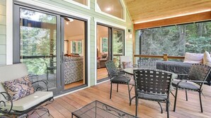 A covered deck with outdoor furniture, large sliding glass doors leading to a bedroom, and a view of the surrounding forest.