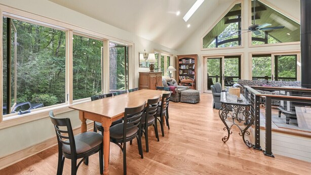 A spacious dining area with a wooden table and chairs, large windows offering a view of the forest, and a high ceiling with skylights.