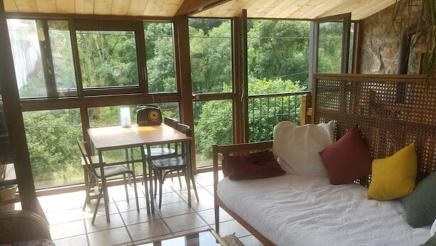 Una habitación con cama, una mesa y sillas. Amplias ventanas ofrecen vistas a árboles.