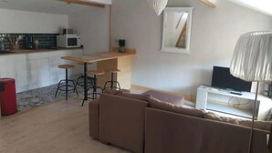 Una sala moderna con un sofá marrón, una mesa de comedor de madera con taburetes de bar, una zona de cocina con microondas y un televisor sobre un mueble blanco.