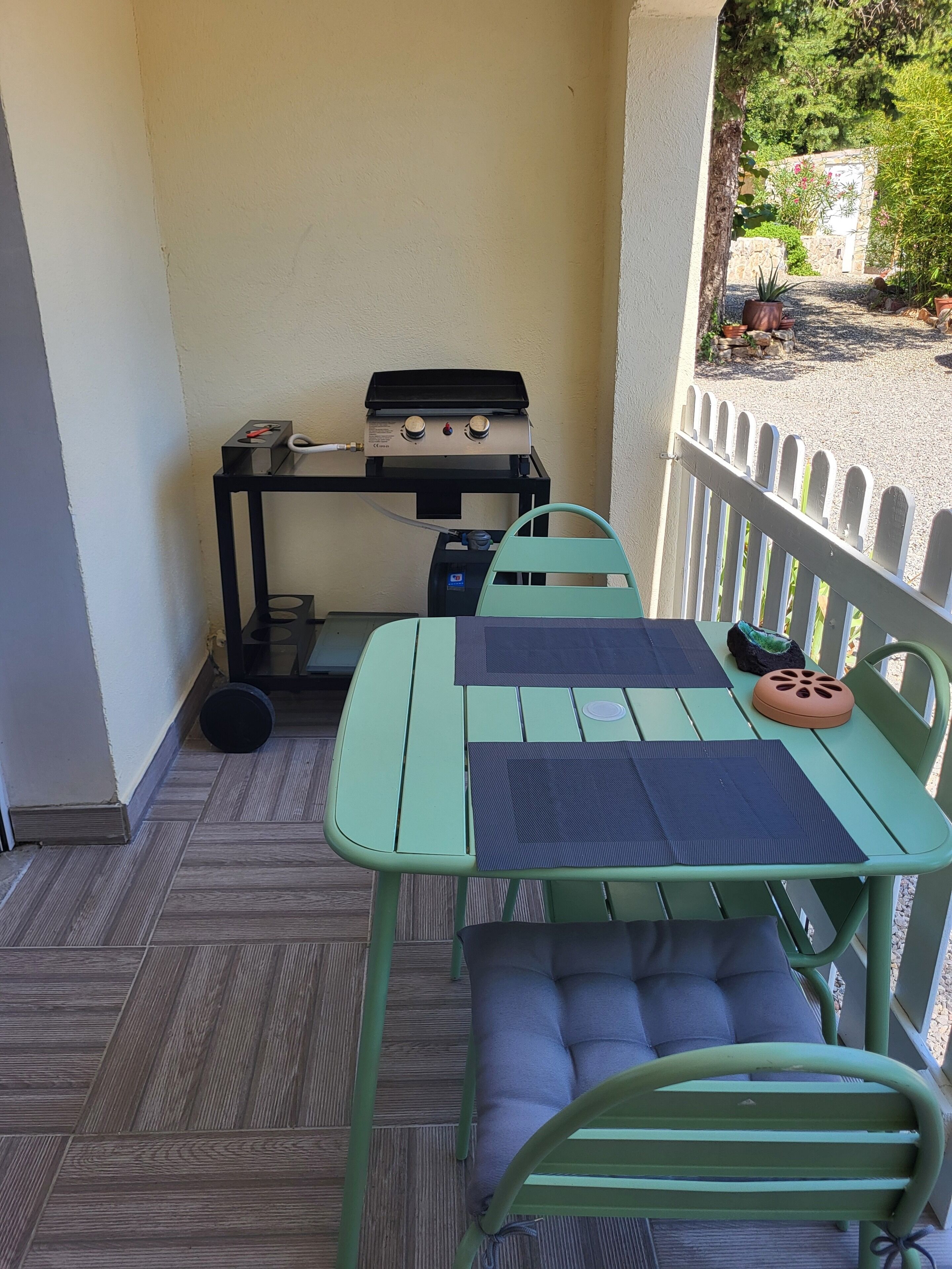 Un petit espace extérieur aménagé en patio, avec une table et des chaises en métal vert, un barbecue et une clôture blanche en bois.