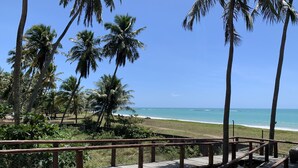 A wooden walkway with a metal railing overlooking a beach with palm trees and a clear blue sky. - Paraíso Frente mar (Maceió)
