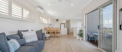 A modern living room with a grey sofa, a dining area with a wooden table, and a kitchen in the background.