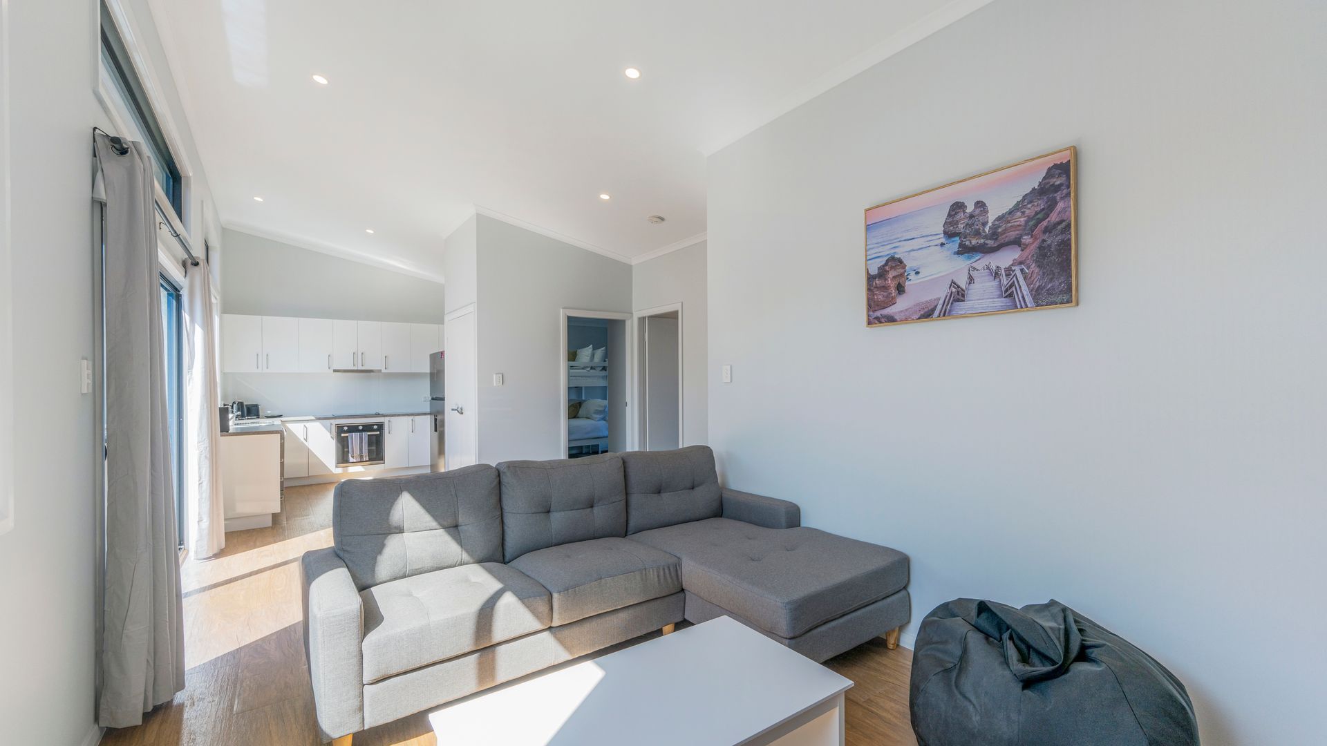 A modern living room with a grey sectional sofa, a white coffee table, and a grey bean bag. There is a kitchen area with white cabinets and a dining table in the background. A painting of a coastal landscape hangs on the wall.