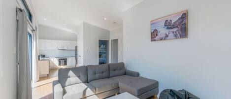 A modern living room with a grey sectional sofa, a white coffee table, and a grey bean bag. There is a kitchen area with white cabinets and a dining table in the background. A painting of a coastal landscape hangs on the wall.