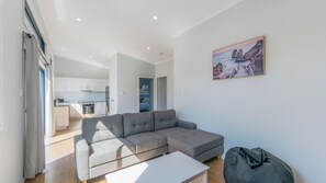 Un salon moderne avec un canapé d’angle gris, une table basse blanche et un fauteuil poire gris. On aperçoit un coin cuisine avec des meubles blancs et, au fond, une table à manger. Un tableau représentant un paysage côtier est accroché au mur.