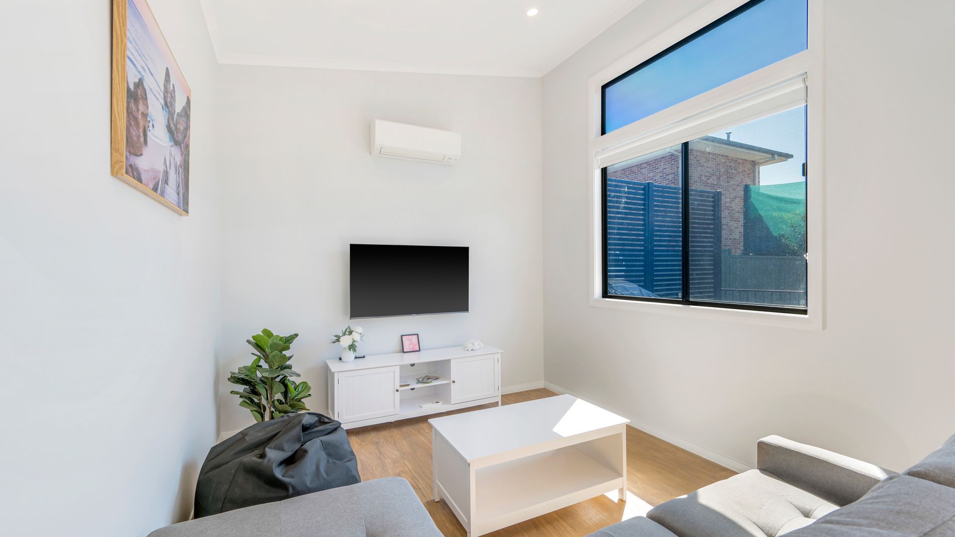 Ein modernes Wohnzimmer mit einem Flachbildfernseher, einer weißen Unterhaltungsanlage, einem grauen Sofa, einem Couchtisch und einem großen Fenster mit Blick auf ein Gebäude und Grünflächen.