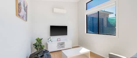 Ein modernes Wohnzimmer mit einem Flachbildfernseher, einer weißen Unterhaltungsanlage, einem grauen Sofa, einem Couchtisch und einem großen Fenster mit Blick auf ein Gebäude und Grünflächen.