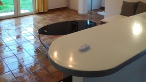A spacious living area with a large sliding glass door leading to a garden, a white sofa, a black chair, and a small table.