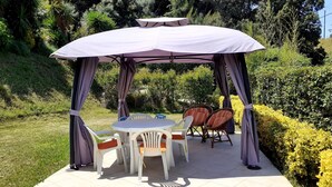 A covered outdoor seating area with a table and chairs, surrounded by greenery.