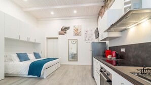 Ein kompakter, moderner Wohnraum mit Küchenzeile, einem Bett mit blauen Kissen und einem großen Spiegel.
