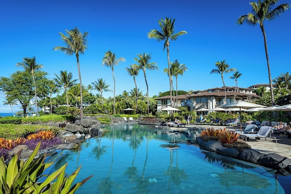 A resort with a clear pool, palm trees, and a building with a beach view.