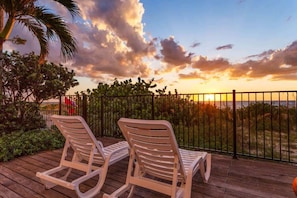 Two white lounge chairs on a wooden deck with a view of the sunset over the ocean.