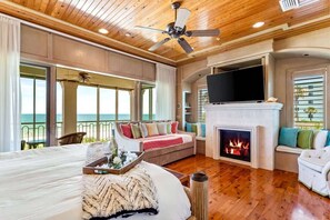 A spacious bedroom with a large bed, a fireplace, a television, and a balcony with a view of the ocean.