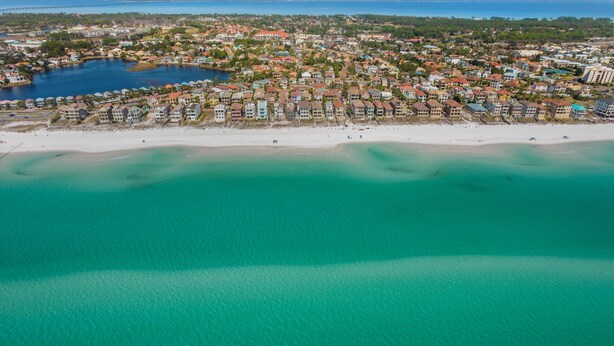 Une vue aérienne d’une ville côtière avec une plage, des eaux turquoise et limpides, et un mélange de bâtiments résidentiels et commerciaux.
