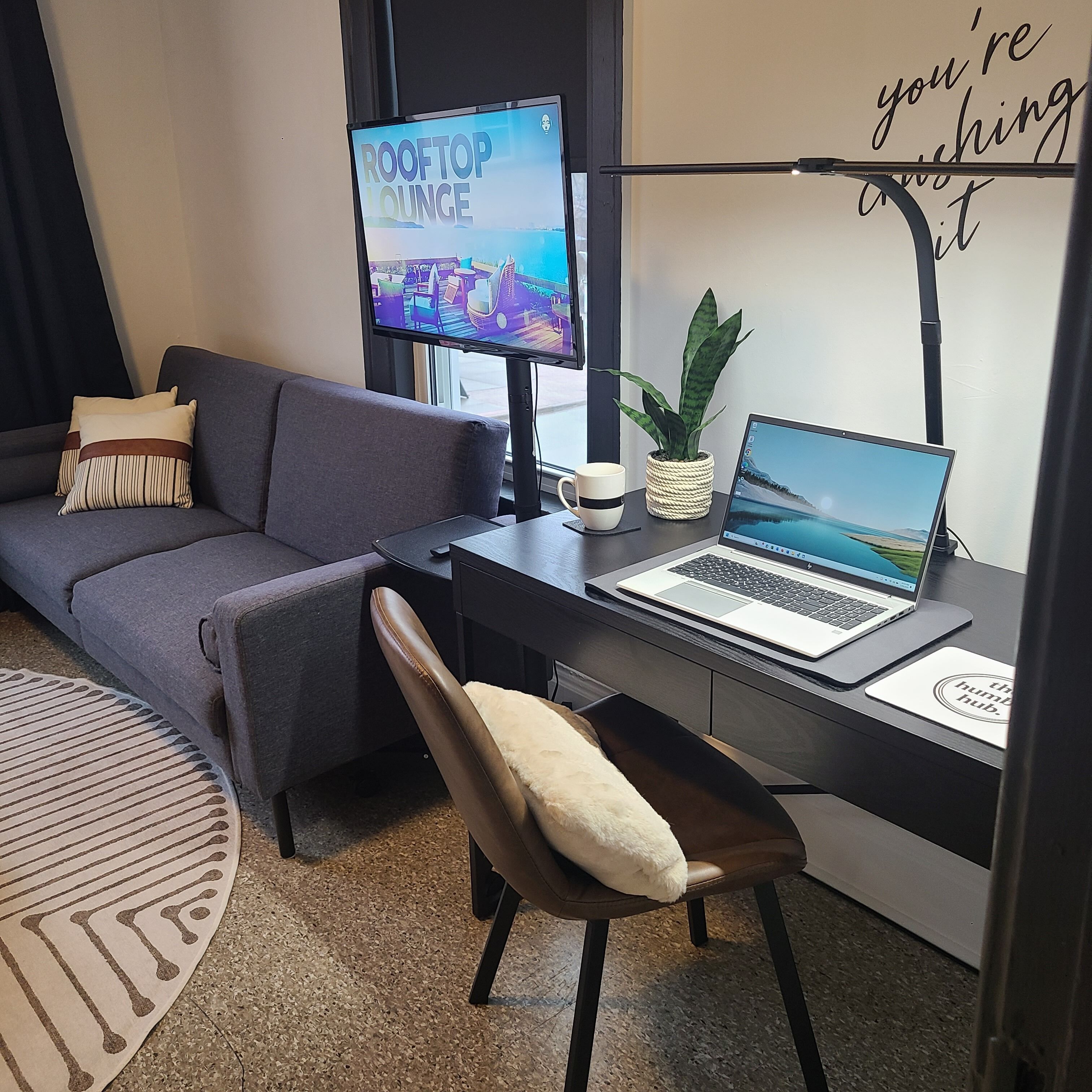 A modern room with a grey sofa, a desk with a laptop, a monitor, a potted plant, and a decorative sign.