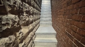 Un escalier étroit, avec des murs en pierre d’un côté et des murs en briques de l’autre, menant à une porte située en haut.