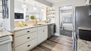 Una cocina moderna con armarios blancos, lavavajillas integrado y un lavamanos. Hay luces colgantes sobre la encimera y una planta pequeña en el alféizar de la ventana.