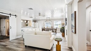 Un salón amplio con un sofá blanco, un comedor con mesa de madera, y una cocina al fondo.