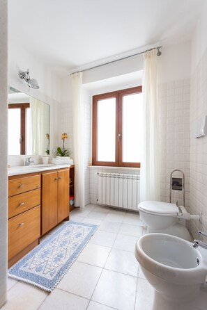 A bathroom with a white sink, wooden cabinets, a blue patterned rug, a toilet, and a window with curtains. - Beautiful and spacious apartment for families in Gallura (Tempio Pausania)