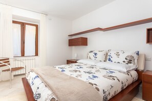 A bedroom with a bed, a nightstand, a wooden bench, a window with curtains, and a built-in shelf above the bed. - Beautiful and spacious apartment for families in Gallura (Tempio Pausania)