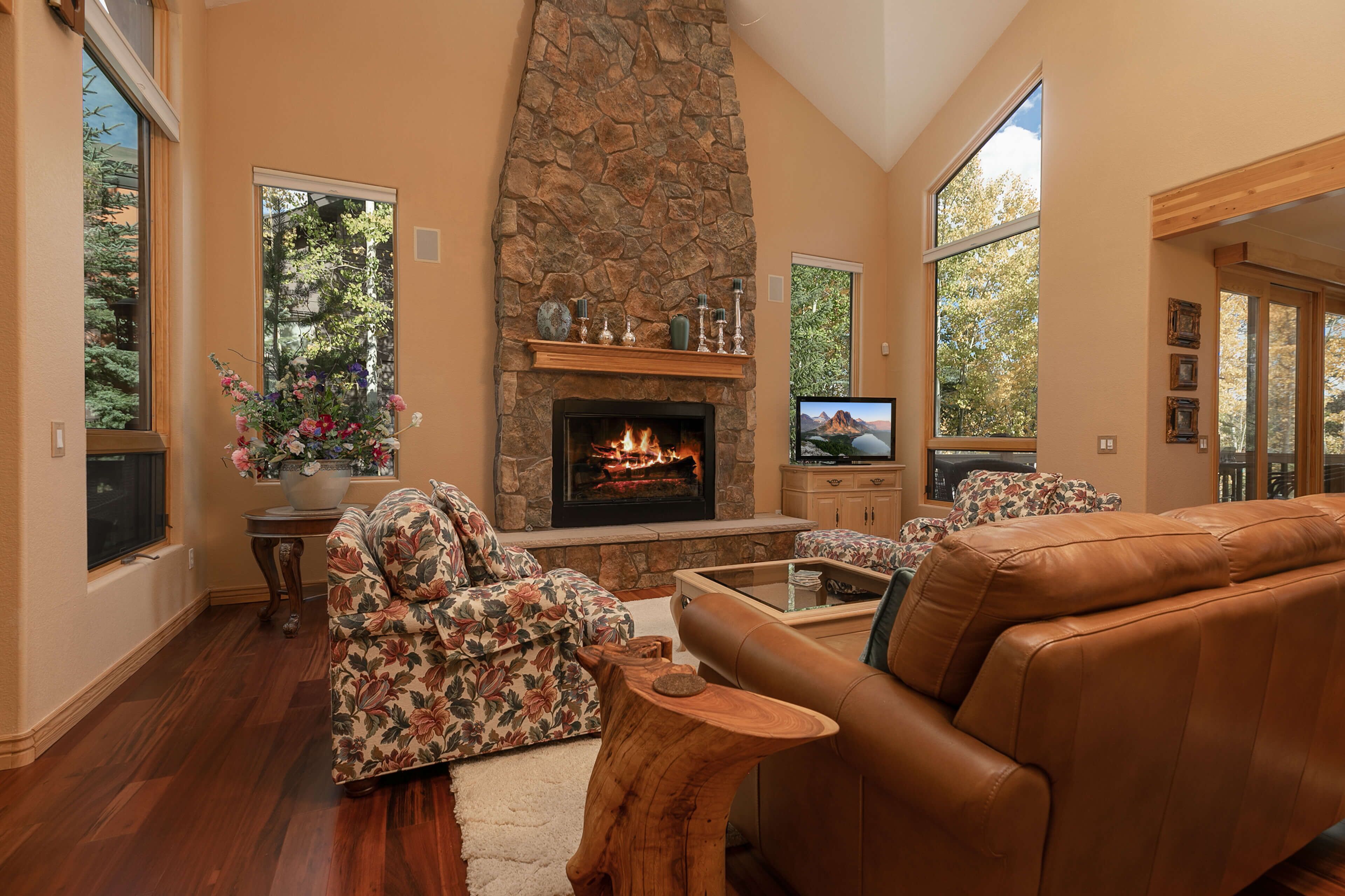 Una sala de estar acogedora con chimenea de piedra, sofá estampado con motivos florales, sofá de cuero y grandes ventanales con vistas a árboles.