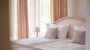 Ein ordentlich bezogenes Bett mit weißen Leinen und Kissen, eine Nachttischlampe und leichte Vorhänge mit Blumenmuster.