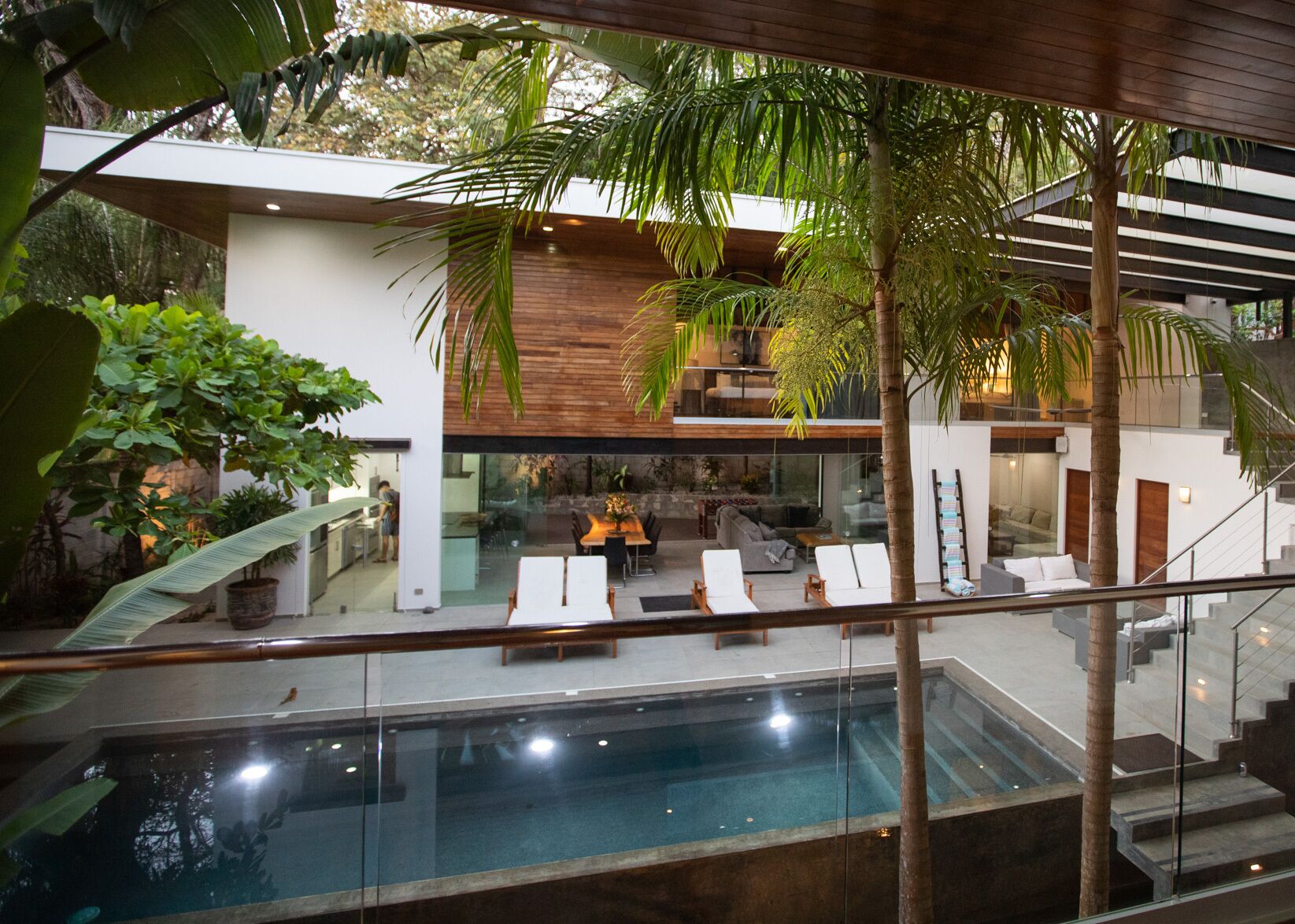 A modern house with a glass-bottomed pool, lounge chairs, and a staircase leading to a second floor.