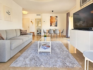 Un salon moderne avec un canapé gris, une table basse sur laquelle sont posés des magazines, un coin repas avec une table et des chaises, et un téléviseur placé sur un meuble.