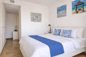 Une chambre à coucher avec un lit, des murs blancs et des œuvres d’art représentant une scène de plage.