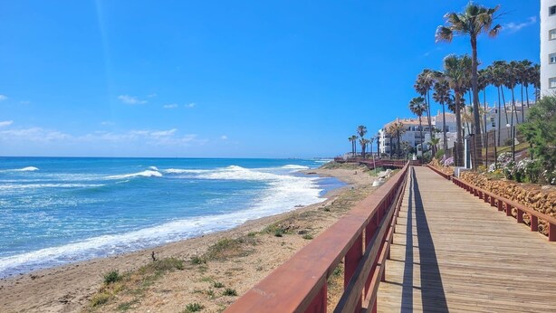 Un muelle de madera a lo largo de una playa, con vistas al océano, palmeras y edificios al fondo.