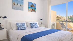 Un dormitorio con una cama grande, ropa de cama blanca, almohadas estampadas en azul y blanco, una manta azul y vistas a un balcón con una silla.