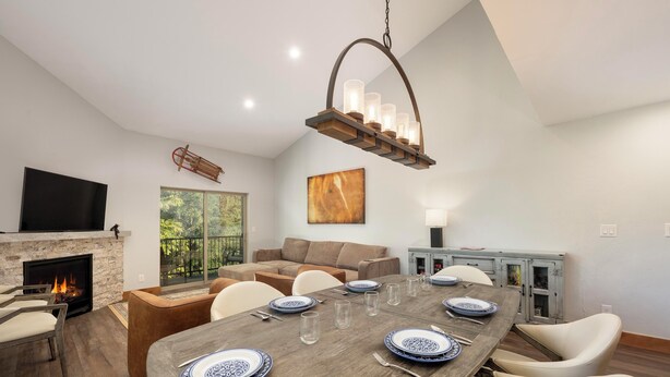 Un coin repas avec une table en bois dressée pour six personnes, une cheminée, un téléviseur et une grande fenêtre donnant sur un paysage verdoyant.