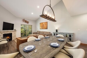 A dining area with a wooden table set for six, a fireplace, a television, and a large window with a view of greenery.