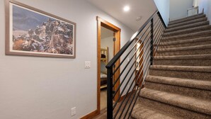 Un escalier dont les marches sont recouvertes de moquette et qui est muni d’une rampe en métal noir, menant à une pièce dotée d’une porte en bois et d’un tableau encadré au mur.