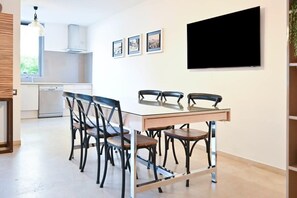 A modern kitchen with a dining area, white cabinets, a large window, and a flat-screen TV mounted on the wall.