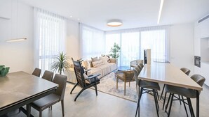 A modern living room with a sofa, dining table, and chairs. There are large windows with sheer curtains, a potted plant, and a ceiling light. - Sweet Home 3BR near the sea by HolyGuest (Tel Aviv-Yafo)