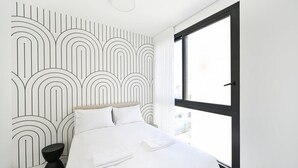 A modern bedroom with a large window, a bed with white linens, and a geometric wall design. - Sweet Home 3BR near the sea by HolyGuest (Tel Aviv-Yafo)