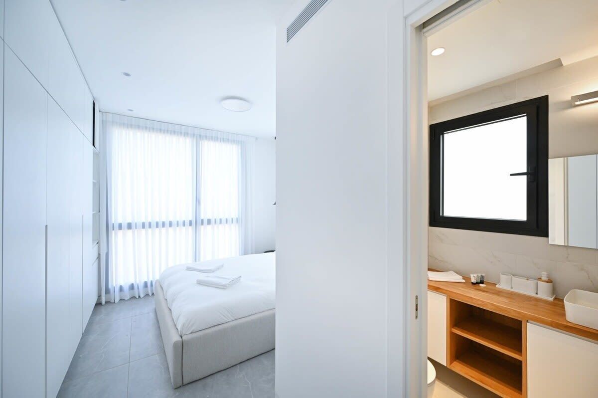 A modern bedroom with a large bed, a wooden vanity, and a large window with blinds.