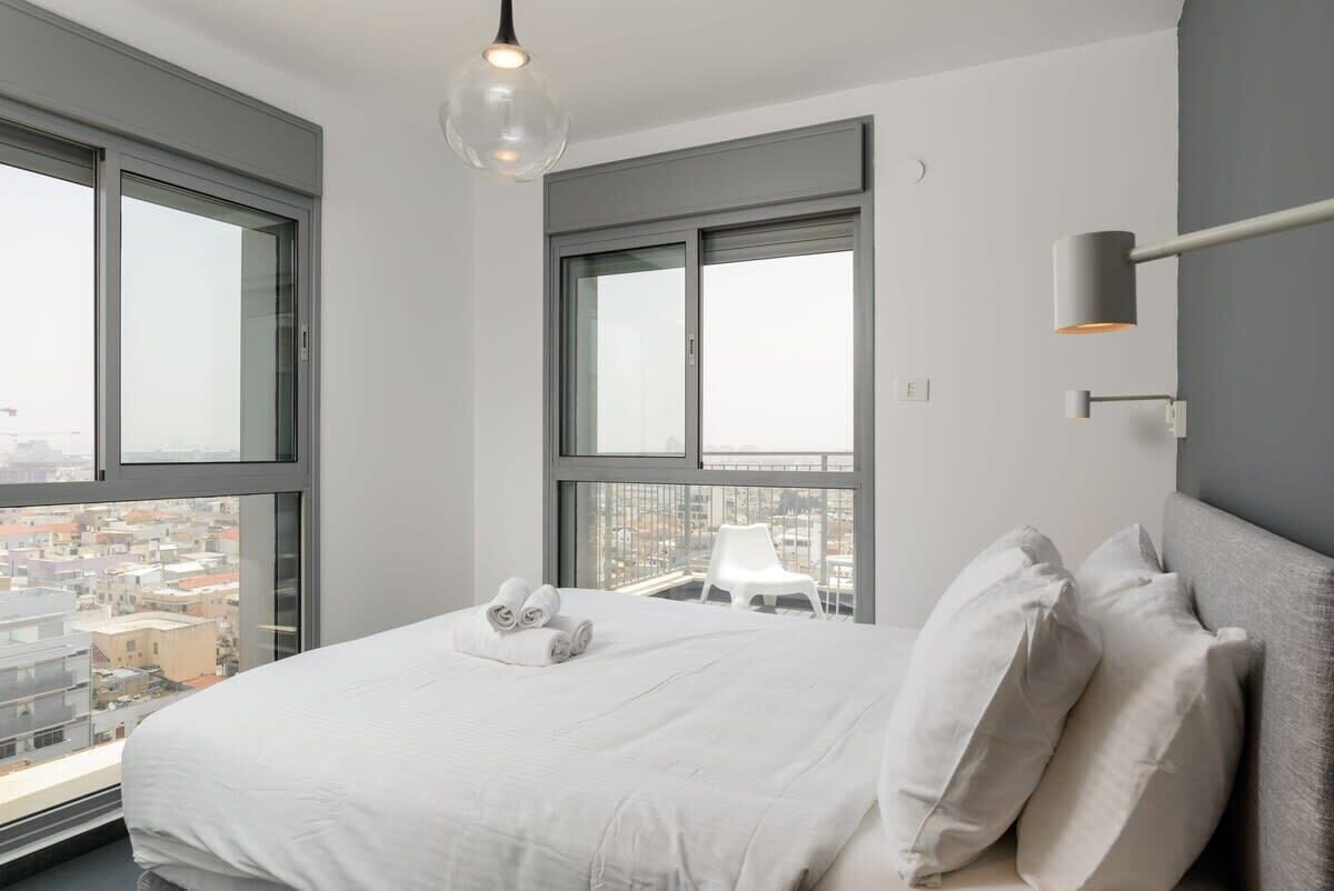 A modern hotel room with a large bed, two windows, a city view, a hanging light fixture, and a side table with a lamp.