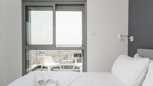 A modern hotel room with a large window, a bed with white linens, and a view of the cityscape.