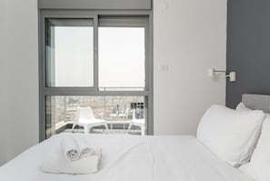 A modern hotel room with a large window, a bed with white linens, and a view of the cityscape.