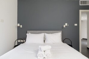 Una cama bien hecha con sábanas blancas y toallas enrolladas, un cabecero gris y dos mesitas de noche con lámparas.