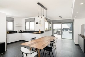 A modern kitchen with a wooden dining table, white cabinets, and a large window offering a view of the city.
