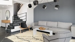 A modern living room with a grey sofa, a black and white patterned chair, a white coffee table, and a staircase with black railings.