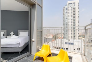 Un dormitorio con cama y mesita de noche, un balcón con sillas amarillas y vistas a un edificio alto.