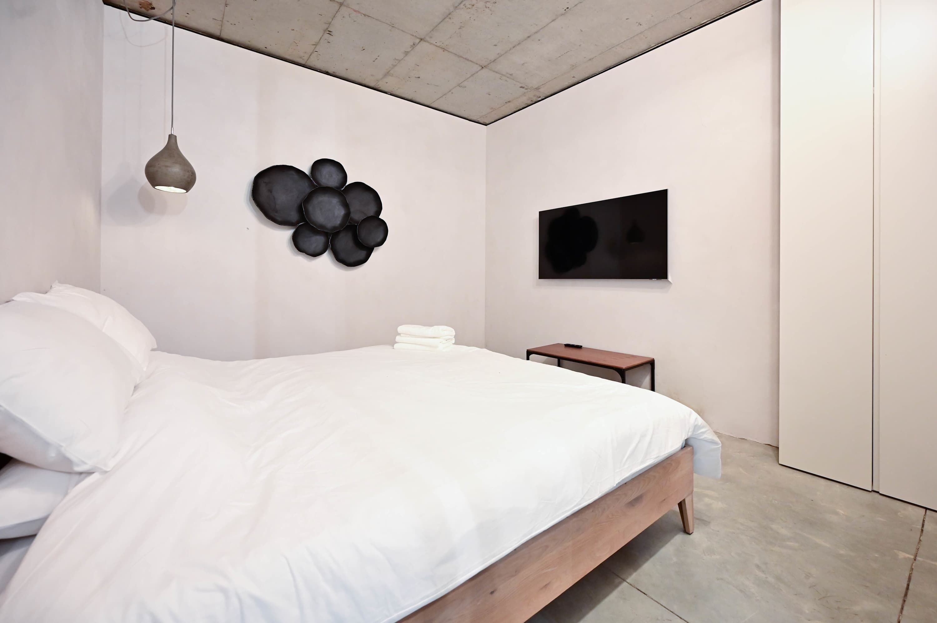 A modern bedroom with a wooden bed, a hanging pendant light, a wall-mounted circular art piece, and a flat-screen TV.